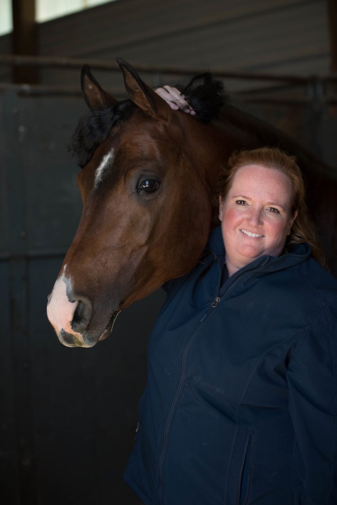Dr. Jennifer Hamilton smiling while posing indoors next to a brown horse in a stable setting.