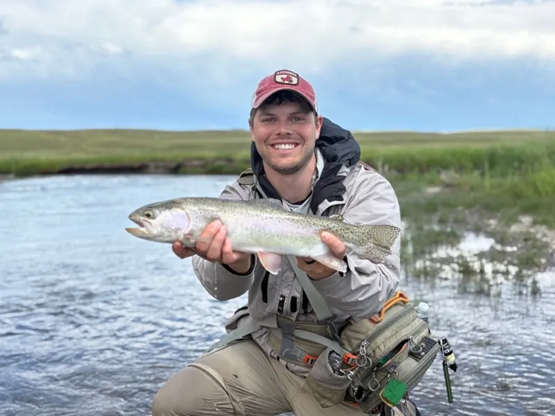 Dr. Isaac Stine smiling outdoors while holding a freshly caught rainbow trout near a river in full fishing gear and a baseball cap.