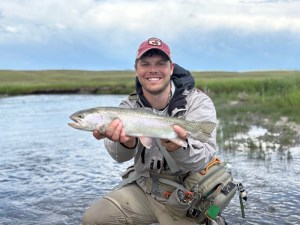 Dr. Isaac Stine smiling outdoors while holding a freshly caught rainbow trout near a river in full fishing gear and a baseball cap.